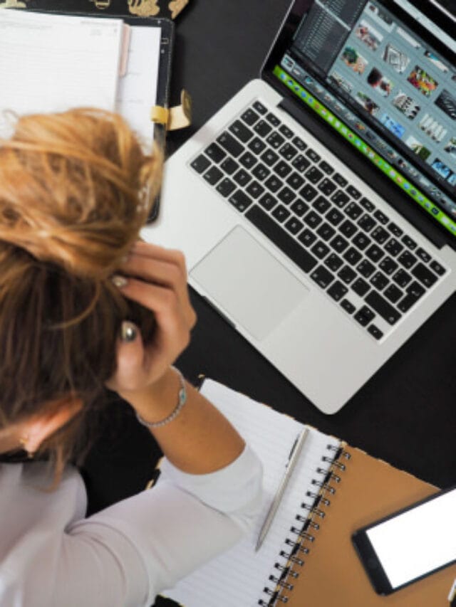 a woman with her head in her hands; she has a messy bun and is sitting in front of a laptop and a notebook